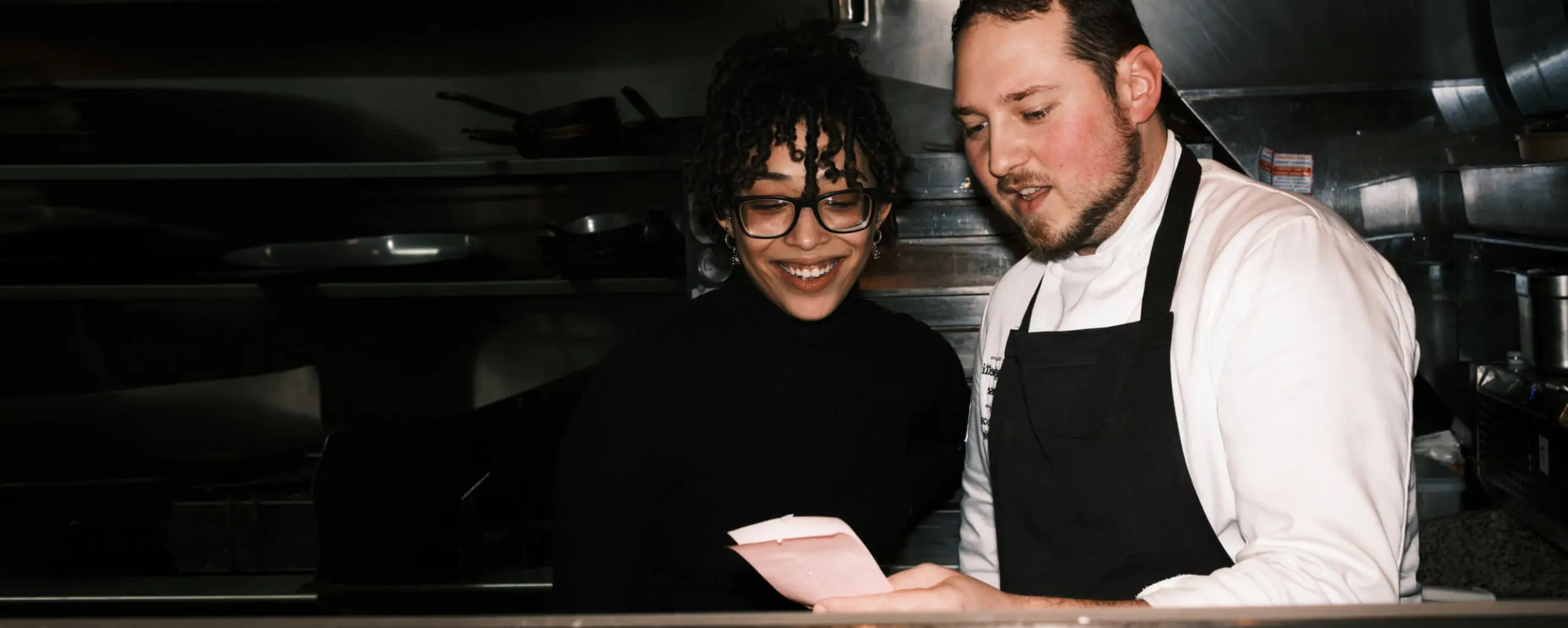 A chef in a white uniform and black apron shows a paper to an Assistant Restaurant Manager with curly hair and glasses in the bustling kitchen. They are both smiling, clearly enjoying their discussion about the latest culinary innovations.