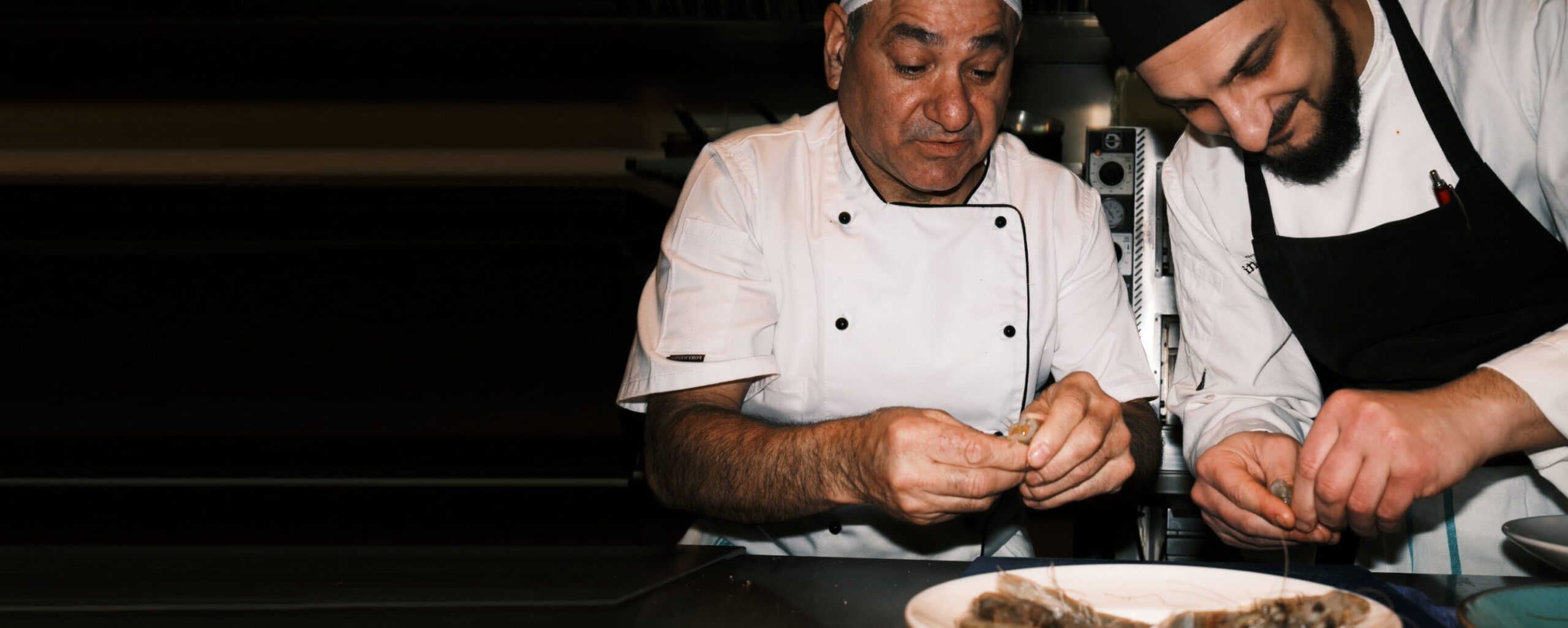 Two chefs in white uniforms are focused on preparing food in a dimly lit kitchen. They are plating a dish, carefully arranging the ingredients on a white plate. The atmosphere is intimate and professional.