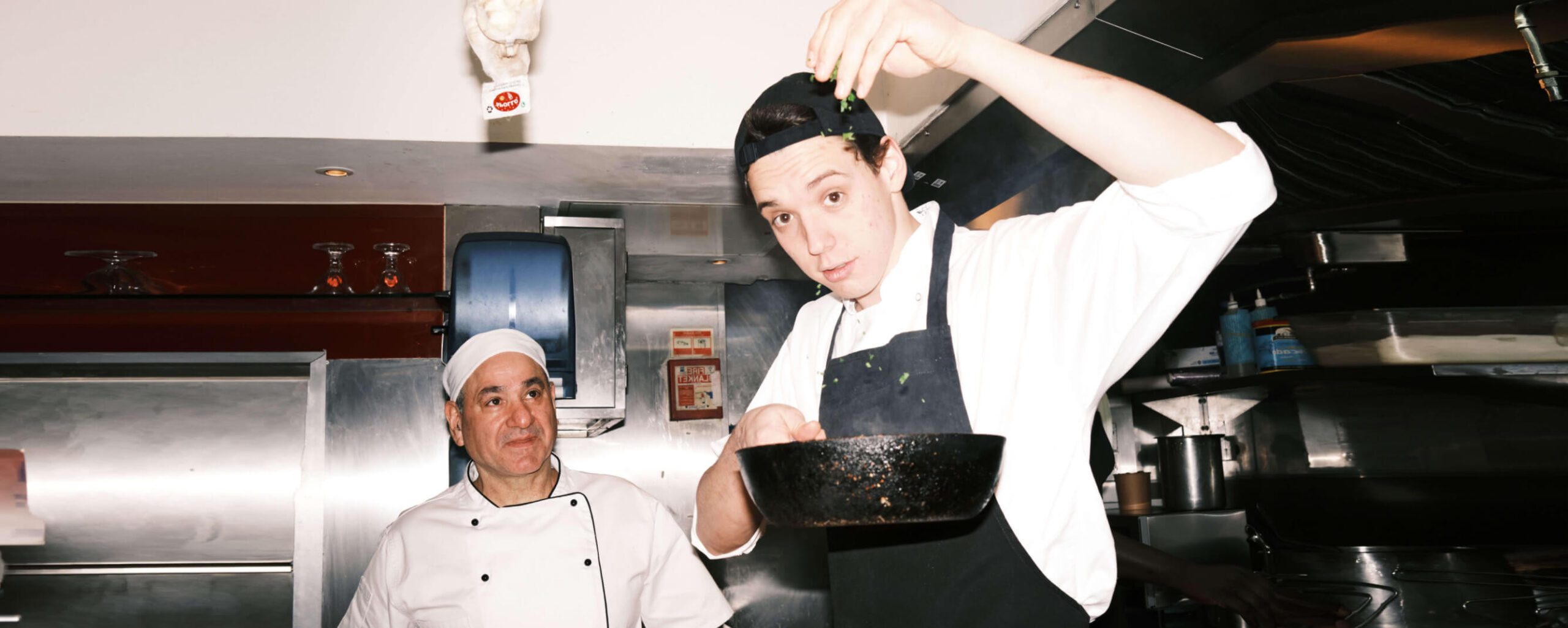 Two chefs in a kitchen: one young chef seasoning a dish in a frying pan, looking focused, wearing a white shirt, apron, and cap. The other chef observes, smiling, wearing a traditional white chef's uniform and hat, with kitchen equipment in the background.