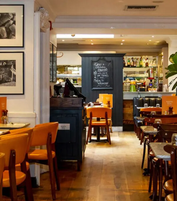 A cozy Italian restaurant on Kensington High Street, with wooden tables and chairs. A blackboard menu hangs near the bar area in the background. Warm lighting fosters an inviting atmosphere, while framed photos adorn the walls.