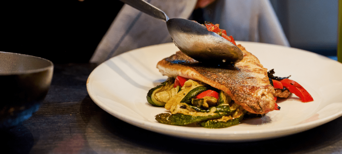 A cooked fish fillet is beautifully arranged on a white plate, served atop roasted zucchini and red peppers. A hand is adding a sauce over the fish with a spoon, enhancing the dish's presentation.