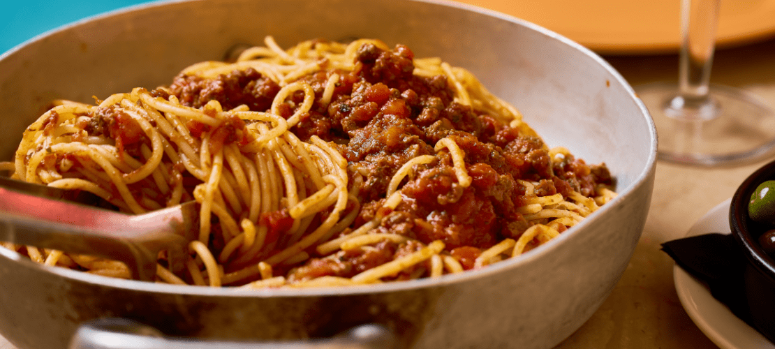 A metal pan filled with spaghetti topped with a rich tomato and meat sauce. A serving fork is partially submerged in the pasta. Nearby, a small bowl contains assorted olives, and there is a blurred glass in the background.
