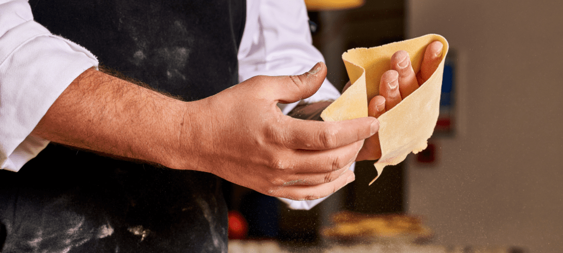 A person wearing a white shirt and dark apron holds a sheet of fresh pasta dough in their hands, with flour dusting the surface. The background is softly blurred, focusing on the pasta preparation process.