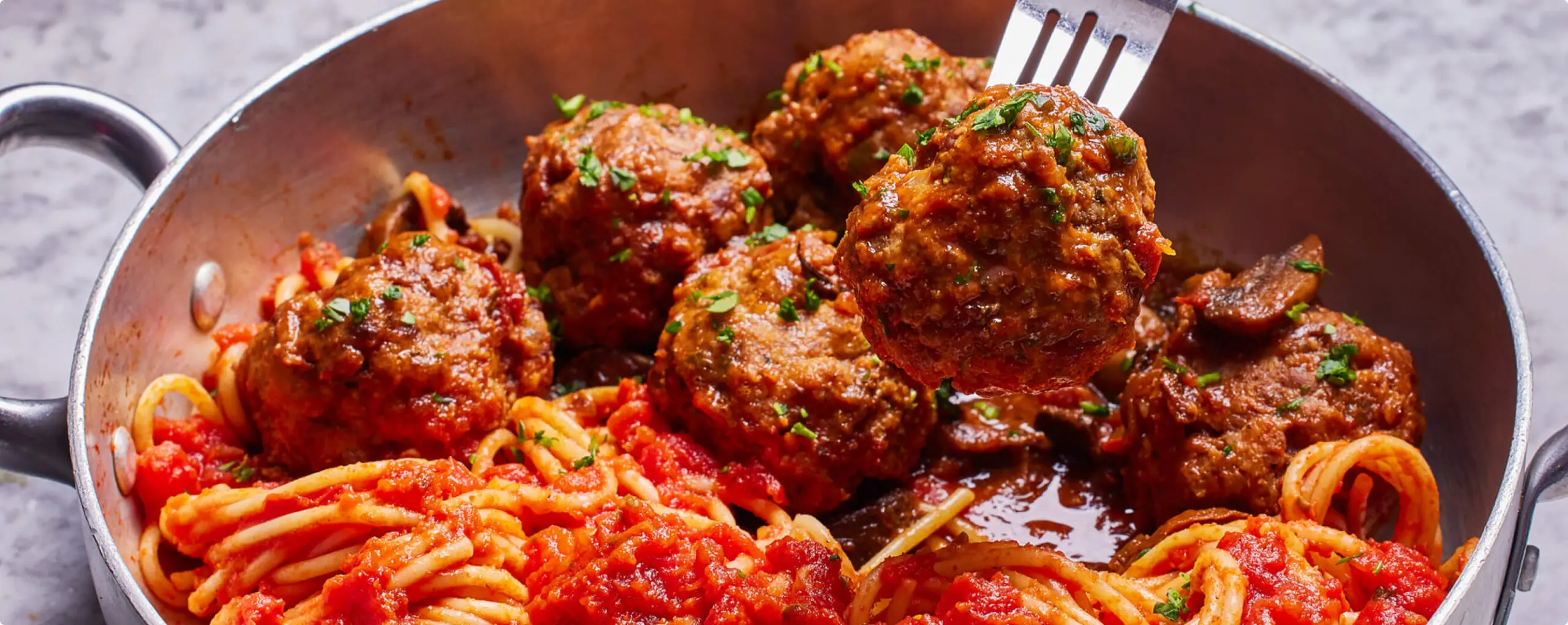 A pan of spaghetti topped with tomato sauce and meatballs. A fork is lifting one of the meatballs, sprinkled with herbs. The dish is set on a gray marble surface.