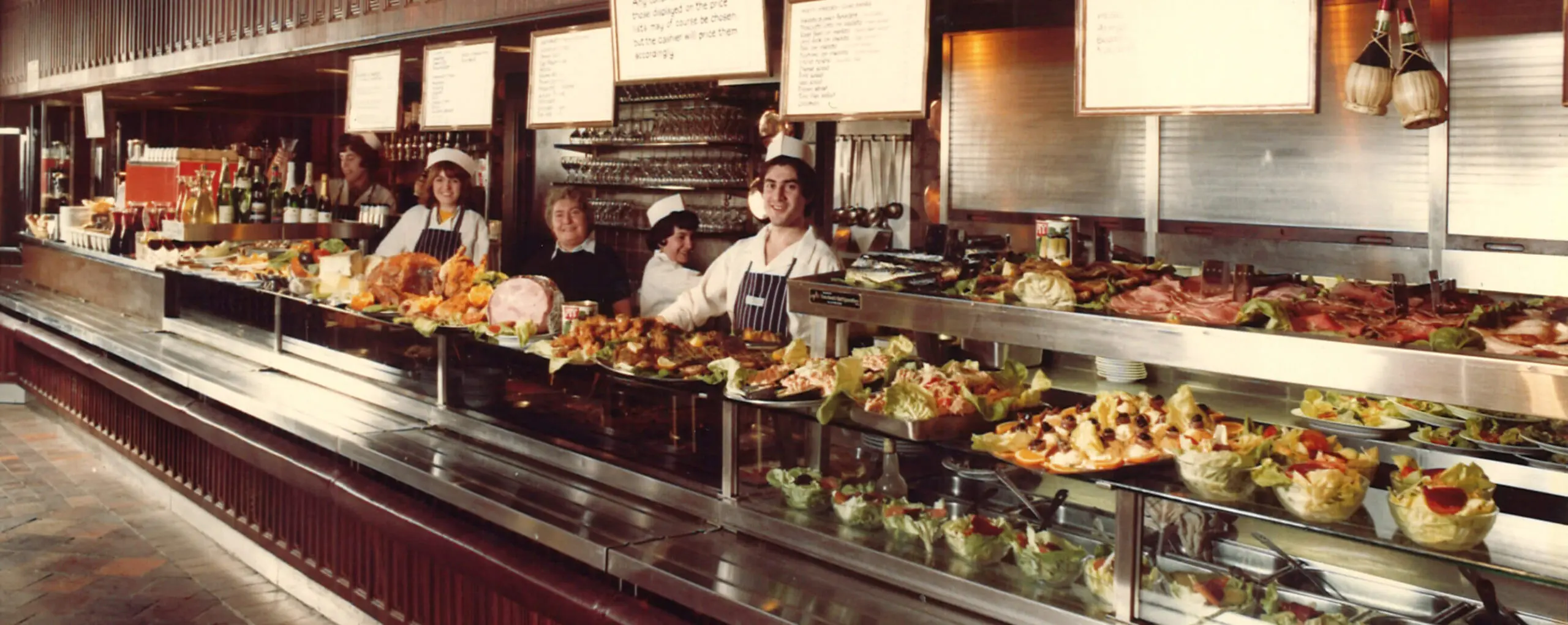 A bustling deli counter with an array of colorful salads, meats, and seafood dishes, displayed in a long, glass case. Several smiling staff members stand behind the counter, ready to serve, with menus hanging above them.
