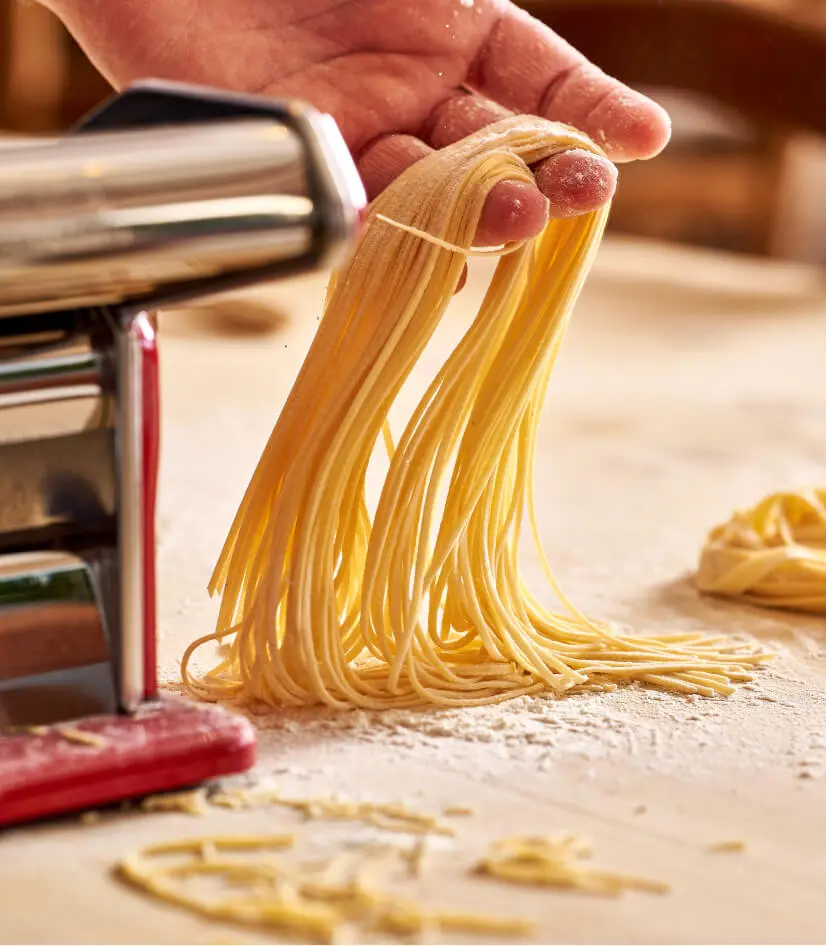 A hand holding freshly made pasta strands next to a pasta machine on a floured wooden surface. The pasta is light in color and has a smooth texture, with more pasta visible in the background.