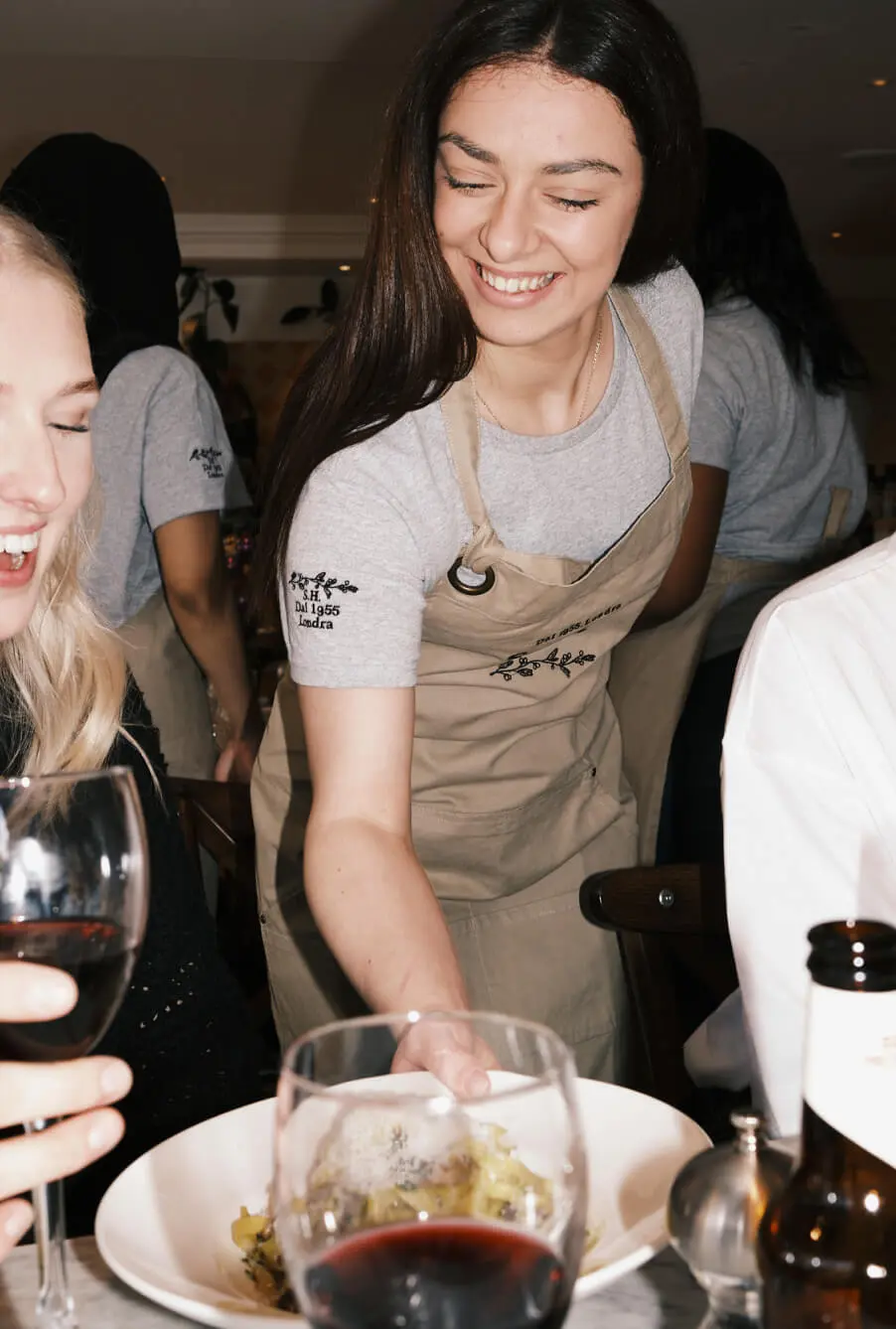 A smiling server in a beige apron places a dish on the table at an Italian restaurant in Westfield White City. Two customers joyfully interact, one holding a glass of red wine, as the lively and warm atmosphere envelops them.