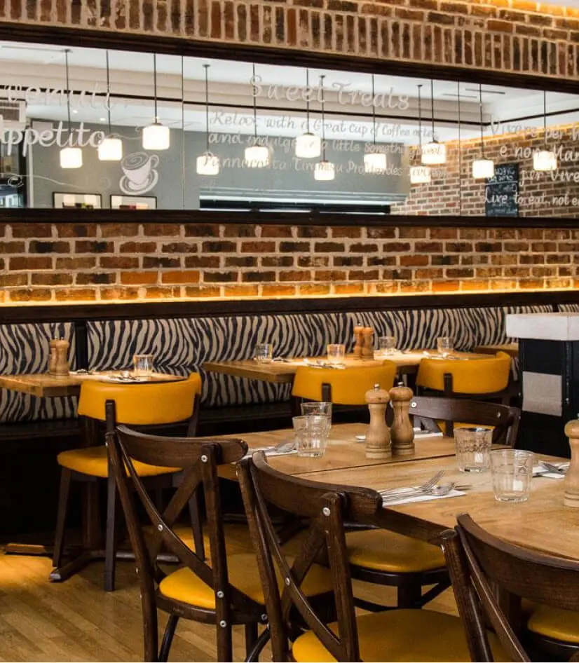 A cozy Italian restaurant on Carnaby Street features wooden tables and chairs, yellow cushioned seating, and exposed brick walls. A large mirror reflects the space, while chalkboard-style writing adorns the walls above. The tables are elegantly set for dining.