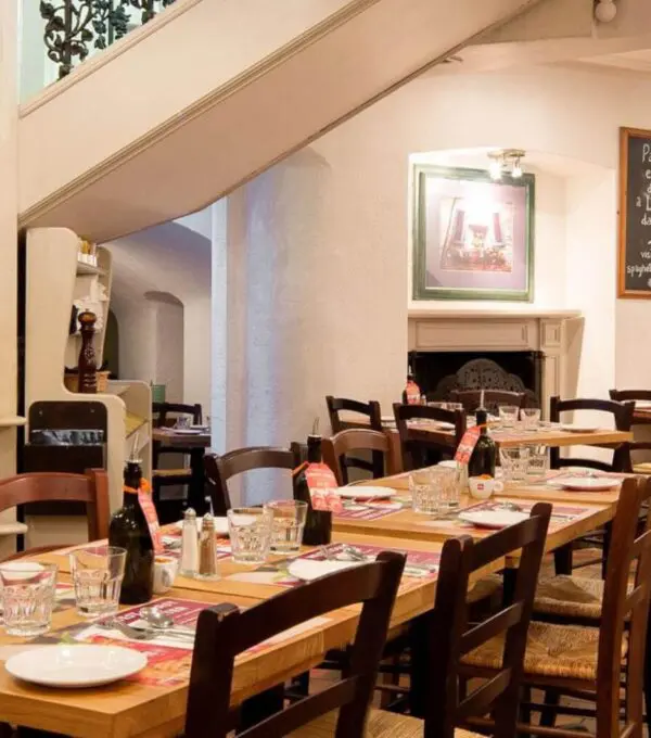 Cozy Italian restaurant interior near Marble Arch, featuring wooden tables and chairs set for dining. The room radiates warmth with its lighting, decorative wall art, and a shelf adorned with wine bottles. On the left, a staircase with an ornate railing adds a touch of elegance.