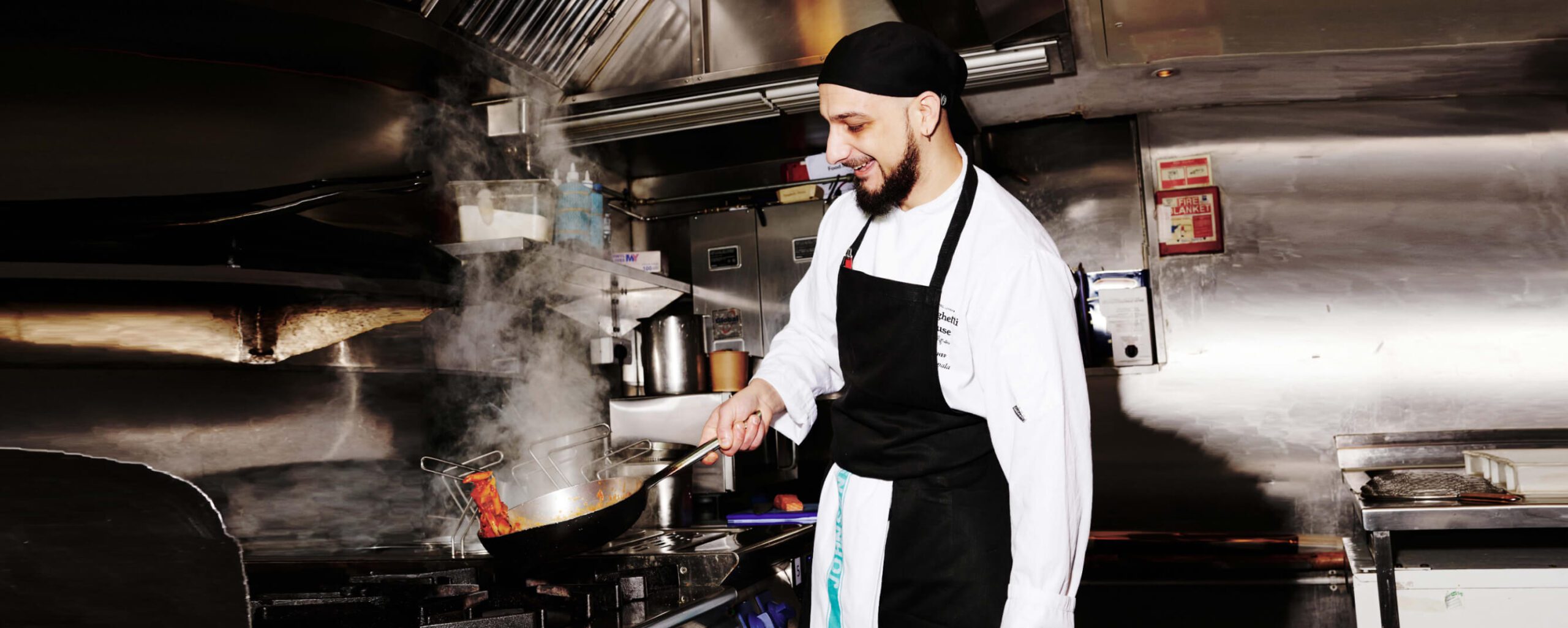 A chef wearing a white uniform and black apron is cooking in a commercial kitchen. He is smiling while flipping food in a pan over a stove with visible steam rising. The kitchen features stainless steel equipment and an oven in the background.
