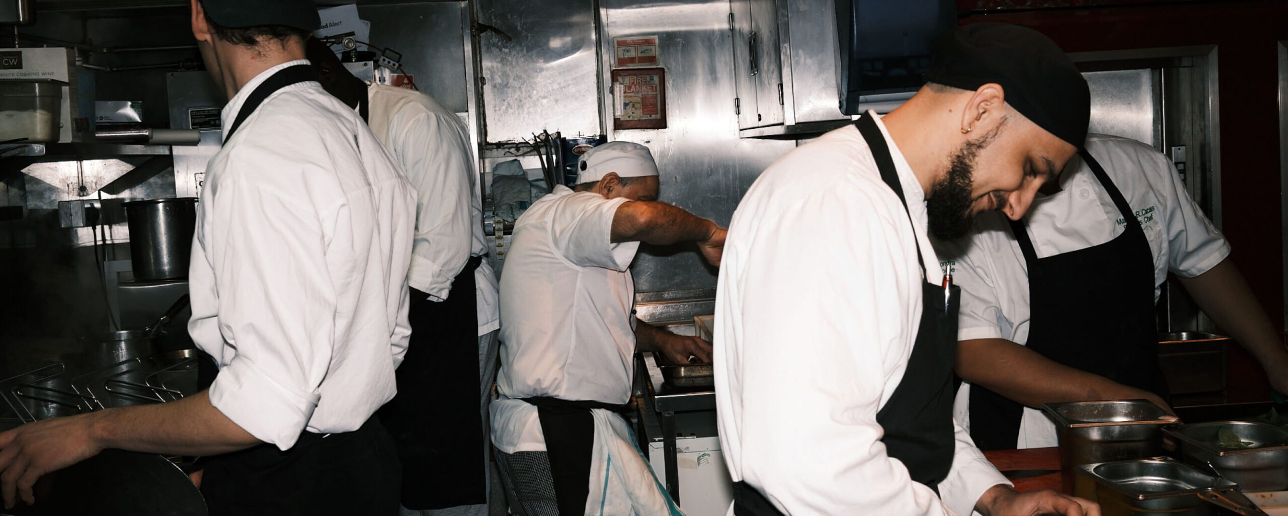 Four chefs in a busy kitchen, all wearing white uniforms and black aprons. One is stirring a pot, while another is focused on preparing a dish. Stainless steel equipment and shelves are in the background, creating a bustling culinary environment.