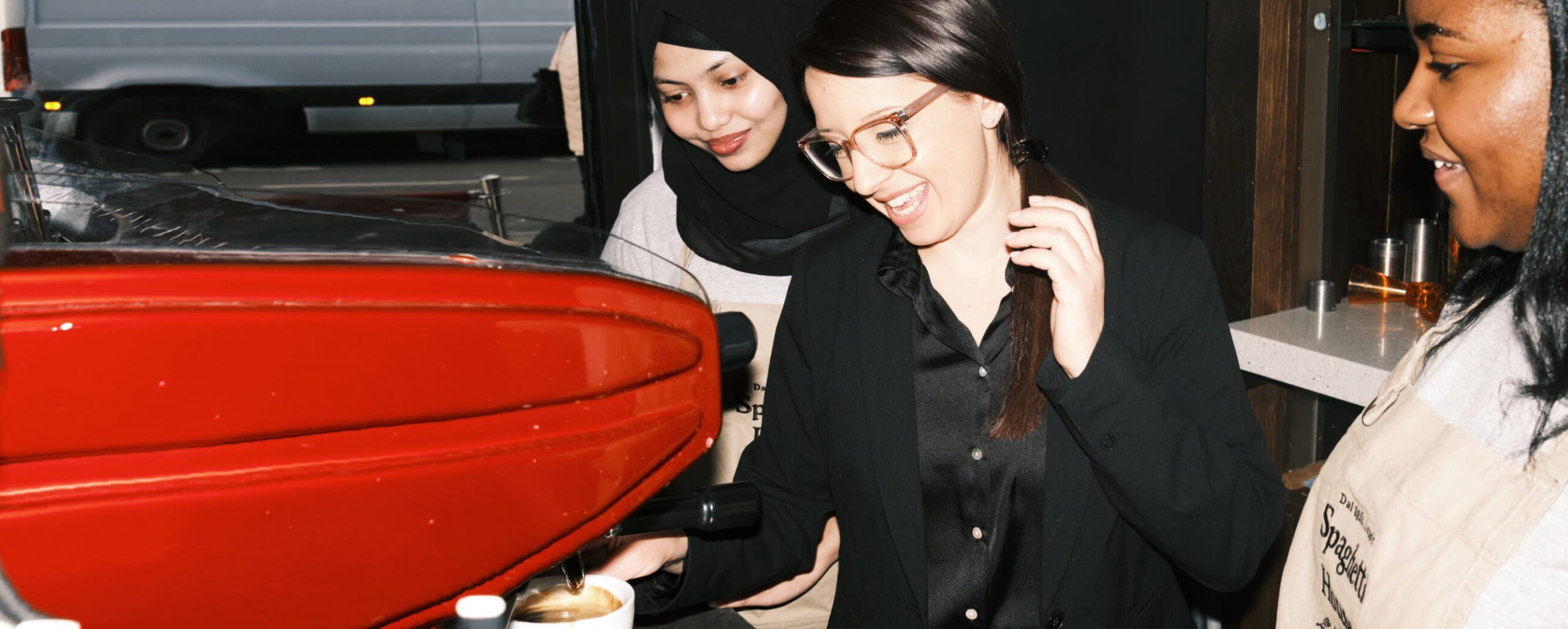 Three people working at a coffee machine. The person in the middle is smiling, wearing glasses and a black shirt. Two others stand beside, one wearing a hijab, both wearing aprons, appearing engaged in a cafe setting.