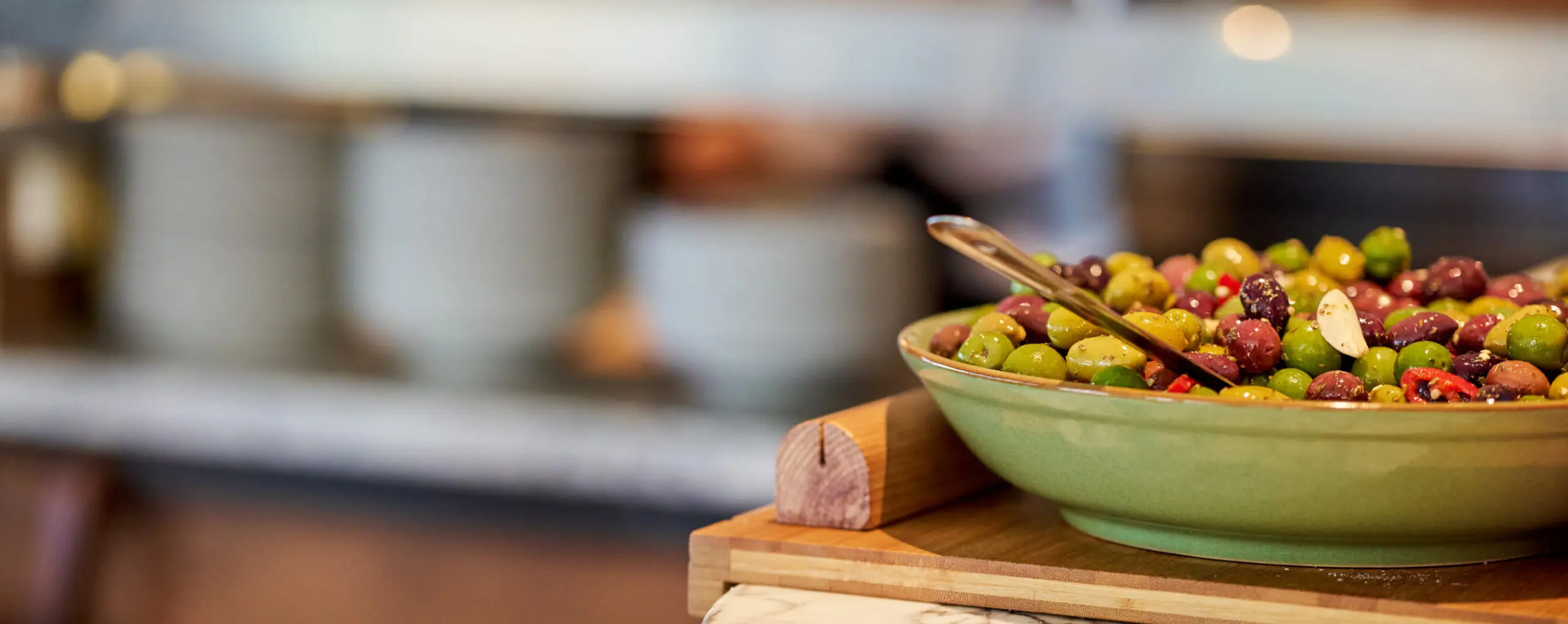 A close-up of a green bowl filled with assorted olives, including green, black, and purple ones, garnished with herbs and garlic slices. The bowl rests on a wooden board with a metal spoon next to it, with a blurred kitchen background.
