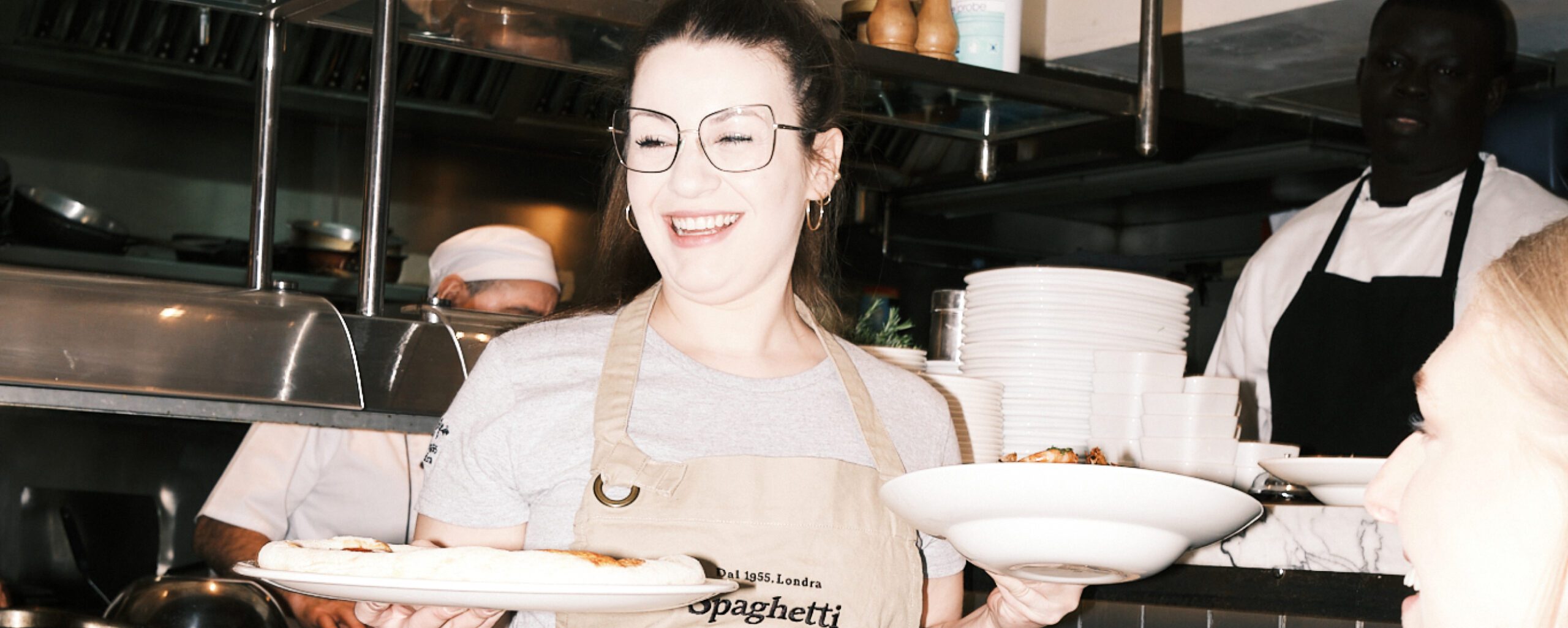 A smiling waitress holds a pizza and a plate of food in a kitchen. She wears an apron and glasses. In the background, a chef and another kitchen staff member are visible. Stacks of plates and kitchen equipment are seen around them.
