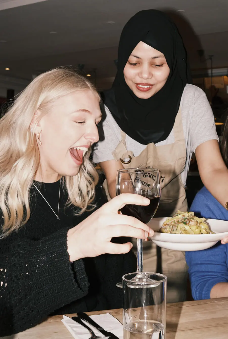 A woman with long blonde hair is smiling and holding a glass of red wine as a server in a hijab and apron brings a plate of pasta to her table at an Italian restaurant in Westfield White City.
