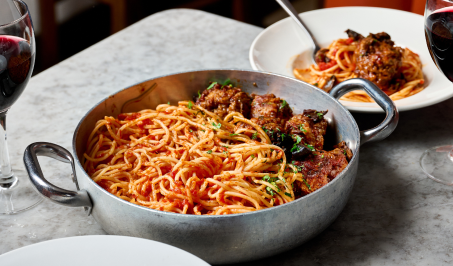A metal pot filled with spaghetti and meatballs garnished with herbs sits on a marble table. In the background, a white plate with a smaller portion and two glasses of red wine are visible, along with a wooden chair.