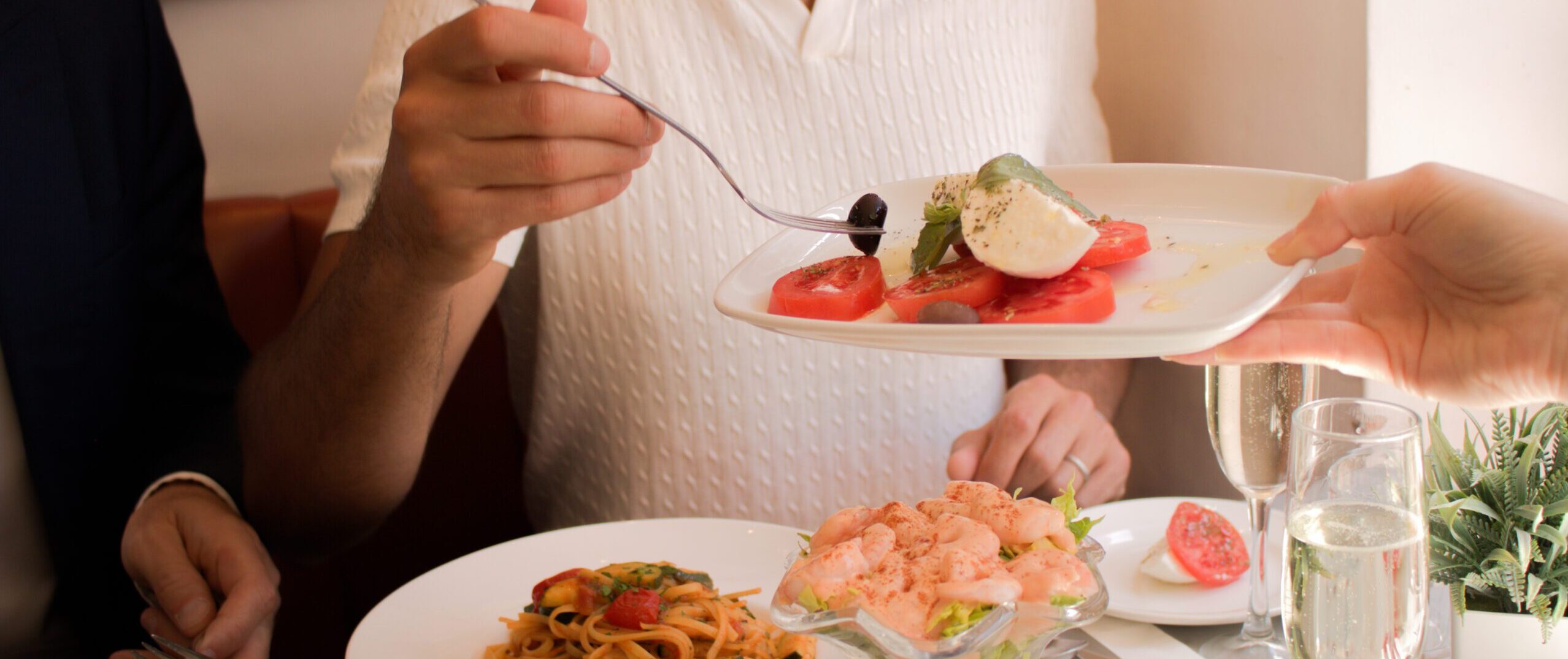 A man in a white shirt is being served a plate of caprese salad with tomatoes, mozzarella, and basil. There are dishes of pasta and shrimp cocktail, and a glass of sparkling drink on the table.